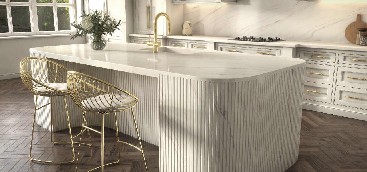 Een elegante keuken met een eiland van Corian® solid surface in een lichte marmerlook. Het aanrecht is omringd door gouden stoelen met een draadframe en beschikt over een gouden kraan.