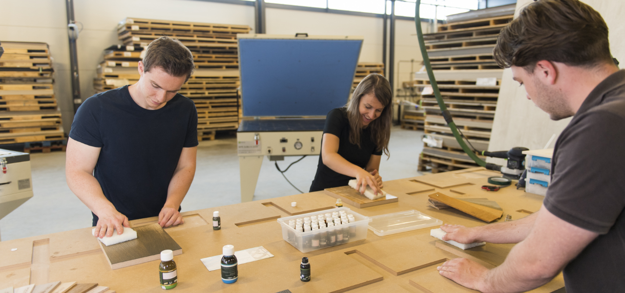 Drie personen in een werkplaats schuren en behandelen houtmonsters op een werkbank in de Leeuwerik Academy fineer, omringd door houten platen en houtbewerking apparatuur.